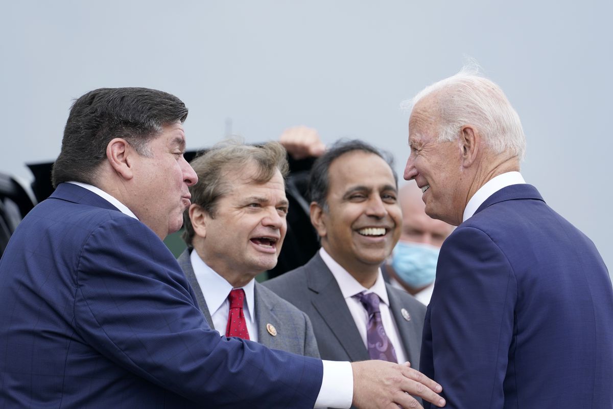 Three men in suits greet each other. On the right is President Joe Biden, smiling, on the left is Governor JB Pritzker, arm outstretched to Biden, in the center is Rep. Mike Quigley, mid  sentence. In the background Rep. Krishnamoorthi is visible.