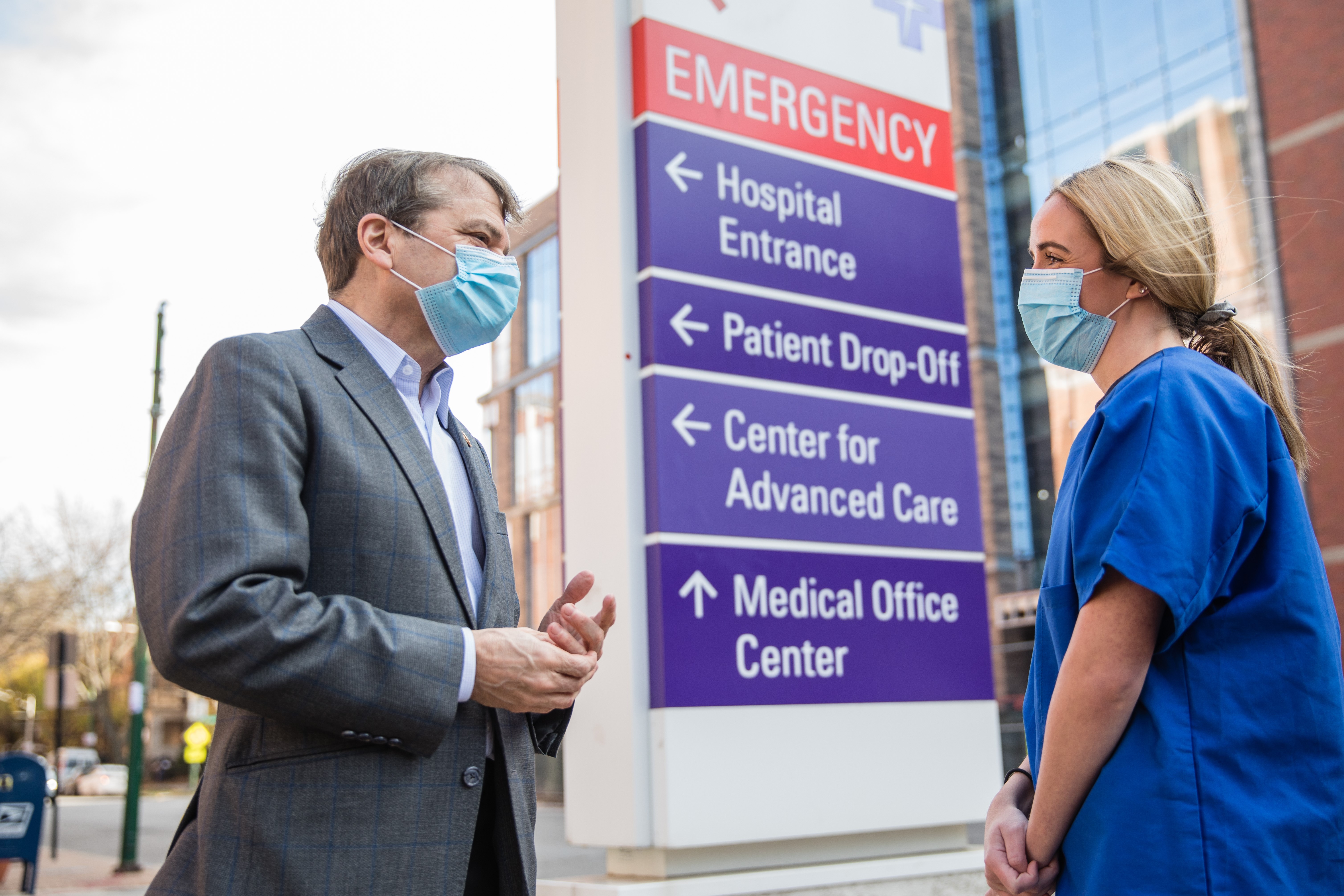 Congressman Mike Quigley wears a suit and facemask speaking with a woman in blue scrubs in front of a COVID testing sign