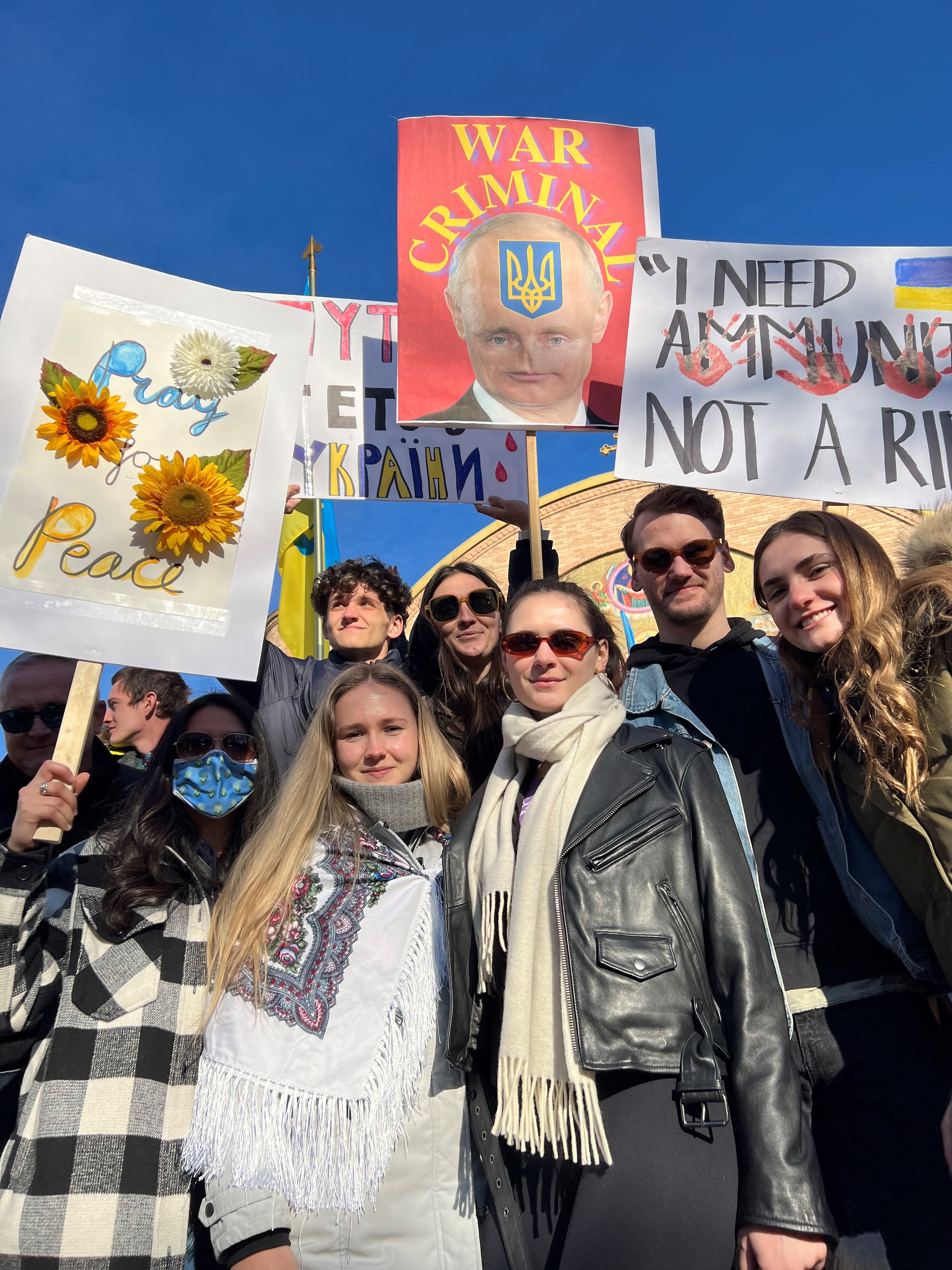 a group of young people stands in a crowd carrying protest signs in support of ukraine