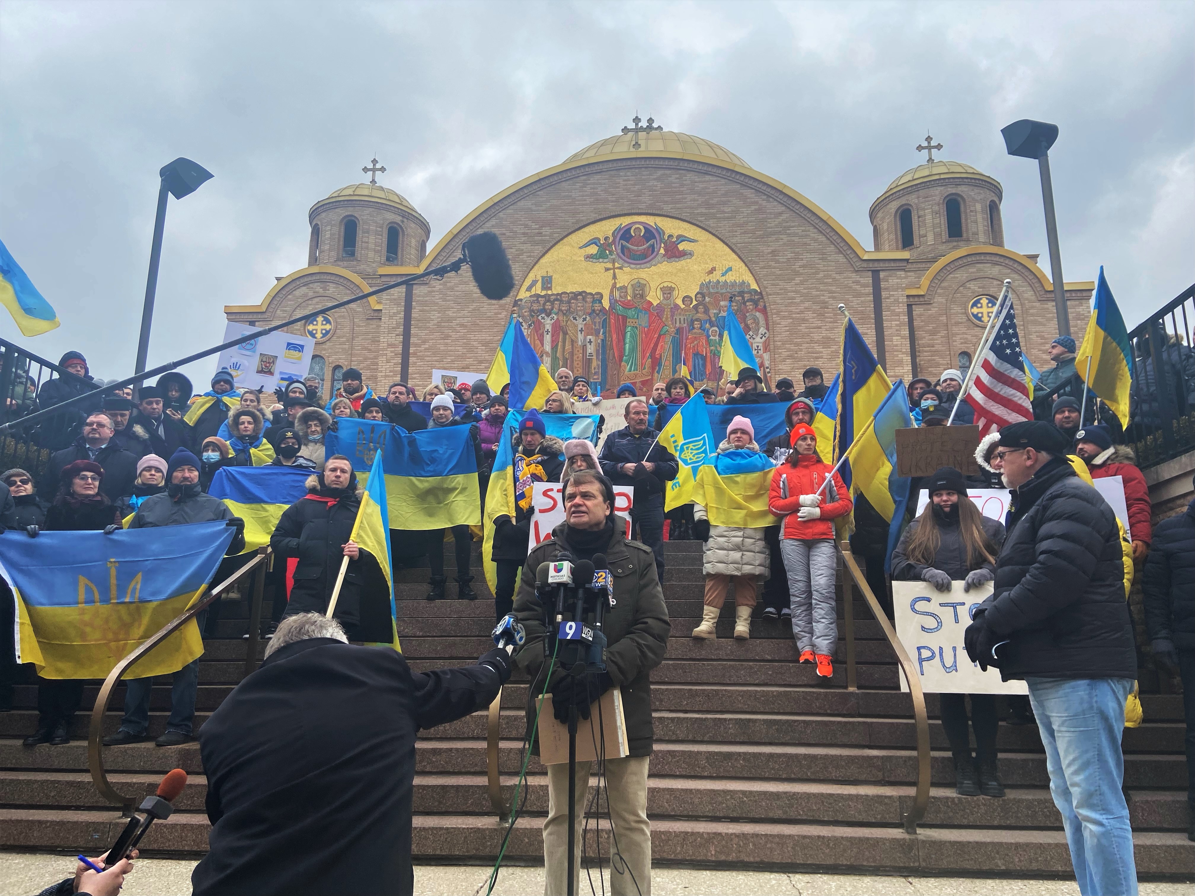 Mike Quigley speaks before microphones, behind him are rally  goers carrying Ukrainian flags