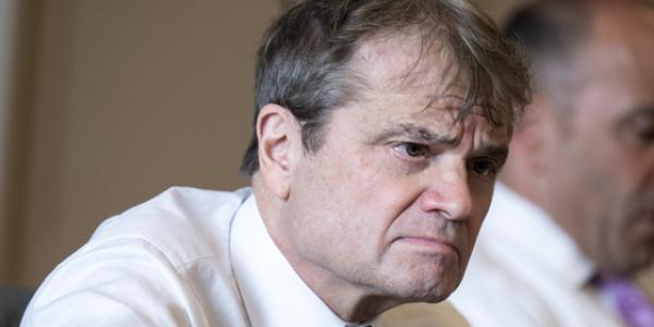 A close up of Quigley wearing a white button down and tie and looking intently at something out of frame