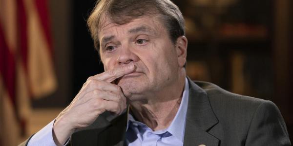 A man in a grey suit in a darkened room with a somber look on his face and his hand to his mouth. The man is Representative Mike Quigley.