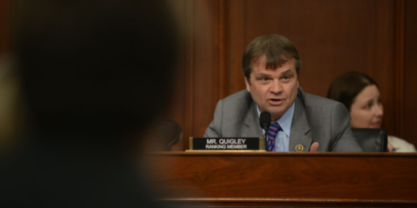 Quigley speaks from a dais in a shirt and tie with a Ranking Member placard in front of him. He appears to be speaking sternly to someone off camera.