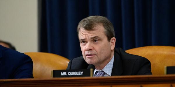 rep quigley sits at a dais with a serious look on his face and a name placard in front of him