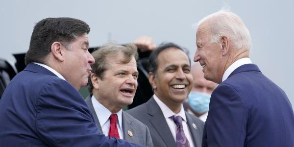 Three men in suits greet each other. On the right is President Joe Biden, smiling, on the left is Governor JB Pritzker, arm outstretched to Biden, in the center is Rep. Mike Quigley, mid  sentence. In the background Rep. Krishnamoorthi is visible.
