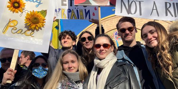 a group of young people stands in a crowd carrying protest signs in support of ukraine