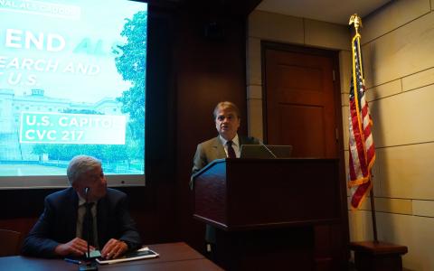 Rep Quigley stands at a podium in a dim room with a brightly lit presentation behind him and a seated panel of experts to his right. 