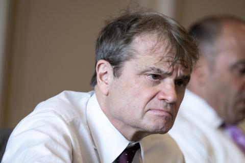 A close up of Quigley wearing a white button down and tie and looking intently at something out of frame
