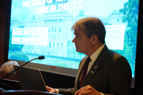 Rep Quigley stands in at a podium in front of a bright blue screen showing a presentation. Quigley wears a suit and is beside a seated panel of people.