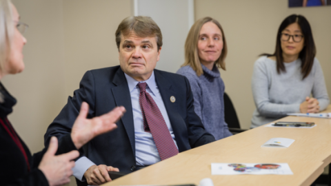 A man in a suit sits at a conference table, turned toward a blonde woman who appears to be addressing him. The man is Rep. Mike Quigley. Two other women are also seated at the table, turned toward Rep. Quigley and the speaker.