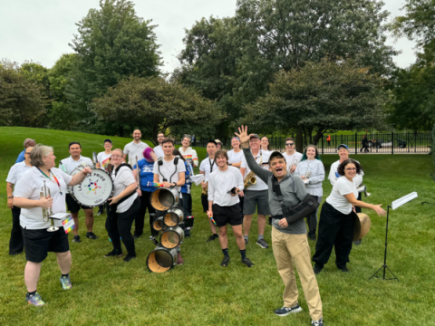 a man in khaki pants and a grey long sleeve with a baseball cap and his left arm in a black sling makes an excited gesture in front of a group of people in matching white t-shirts and carrying instruments, clearly a marching band. the man is mike quigley.