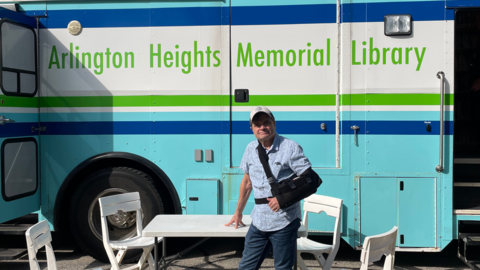 a man in a button down and black sling stands in front of a white, blue, and green bus that has signage labeling it the arlington heights memorial library