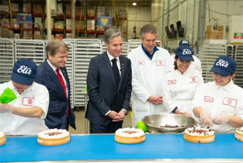 mike quigley stands in front of a cheesecake conveyor belt surrounded by men and women in white eli's cheesecake lab coats.