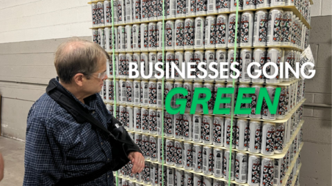 a man wearing a blue flannel and safety goggles with his left arm in a bulky sling looks at a large stack of beer cans