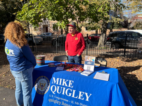 A man in a red shirt and a Cubs baseball cap stands behind a table with literature on it and a blue table cloth that says "Mike Quigley". The man is Mike Quigley. He is talking to a woman who stands on the other side of the table. They are in a park with trees behind them.