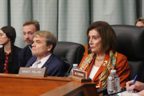 an older woman with short brown hair, an orange blazer, and a colorful scarf sits at a lectern beside a man in a blue suit. both are staring straight ahead with serious, somber looks on their faces. the woman is nancy pelosi and the man is mike quigley.