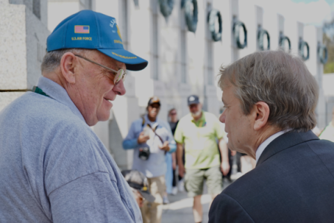 You see Congressman Mike Quigley in a suit from the back, speaking with a man in a grey polo and blue baseball cap. His cap indicates that he is a veteran. In the background is a the World War II Memorial in Washington, DC