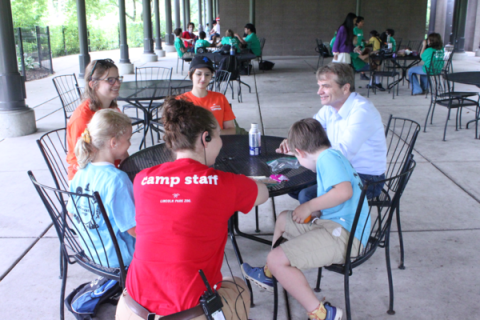 Mike Quigley sits at a metal table with two second graders in blue shirts and two teenagers in orange shirts. A camp staff member in a red shirt kneels beside the table. All are smiling.
