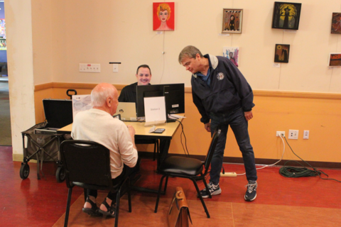 a man in a windbreaker leans over two men seated at a small table. the man in the windbreaker is rep. mike quigley. the two seated men are a veteran and a VA representative.