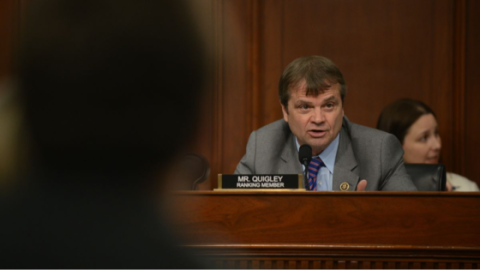 Quigley speaks from a dais in a shirt and tie with a Ranking Member placard in front of him. He appears to be speaking sternly to someone off camera.