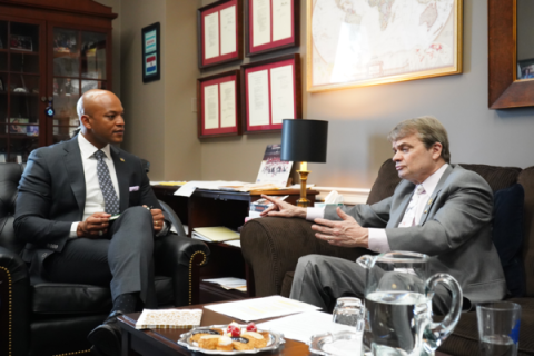 Rep. Mike Quigley sits on a brown couch in an office. He is gesticulating while speaking to a man seated diagonally from him. Both wear nice suits. The other man is Governor Wes Moore.