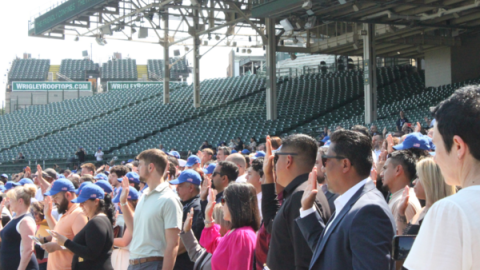 hundreds of individuals in Cubs baseball caps stand with their right hands raise in the stands of Wrigley Field