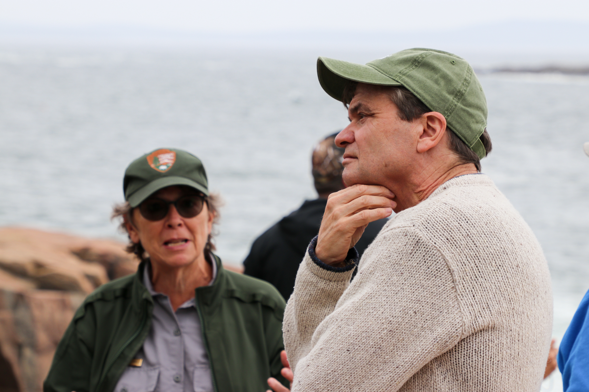In the foreground, Congressman Mike Quigley stands with his arms crossed as he listens to a woman in a green national park service hat. The ocean is in the background.