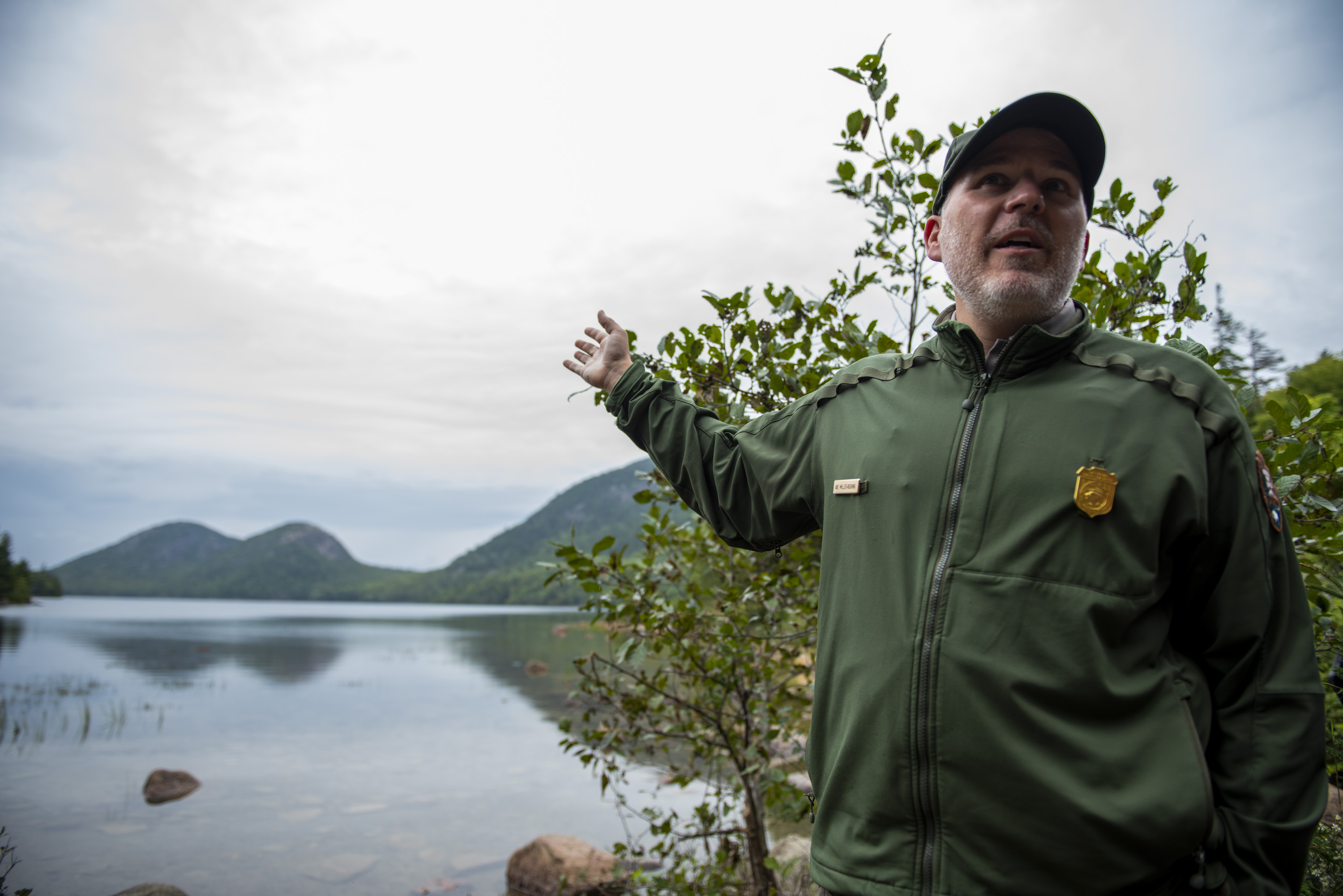 A man in a National Park Service uniform and badge speaks and gestures toward a lake with green hills in the background.