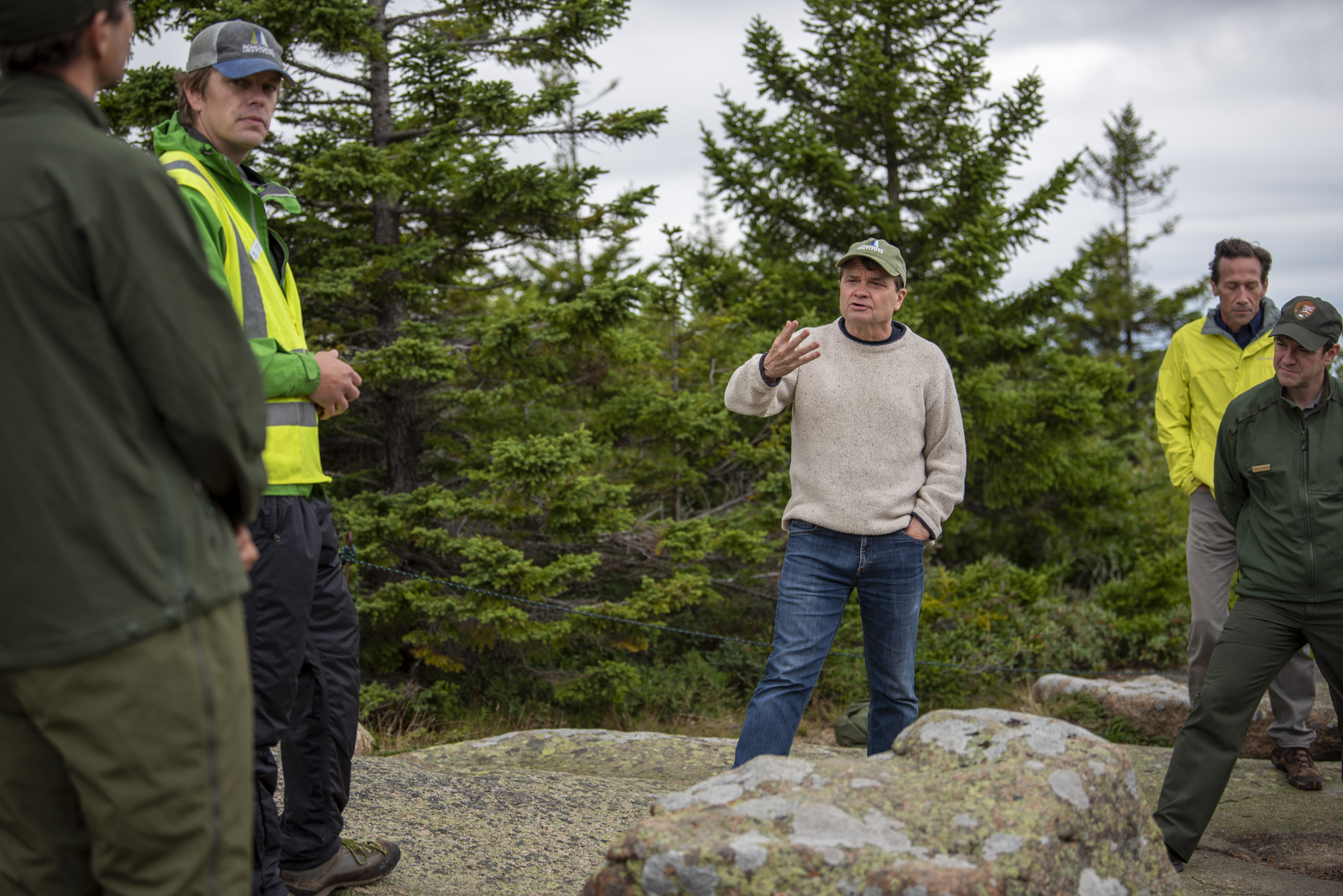 Congressman Mike Quigley stands in front of a line of trees and gesticulates as he speaks to men wearing yellow safety vests and green National Park Service gear.