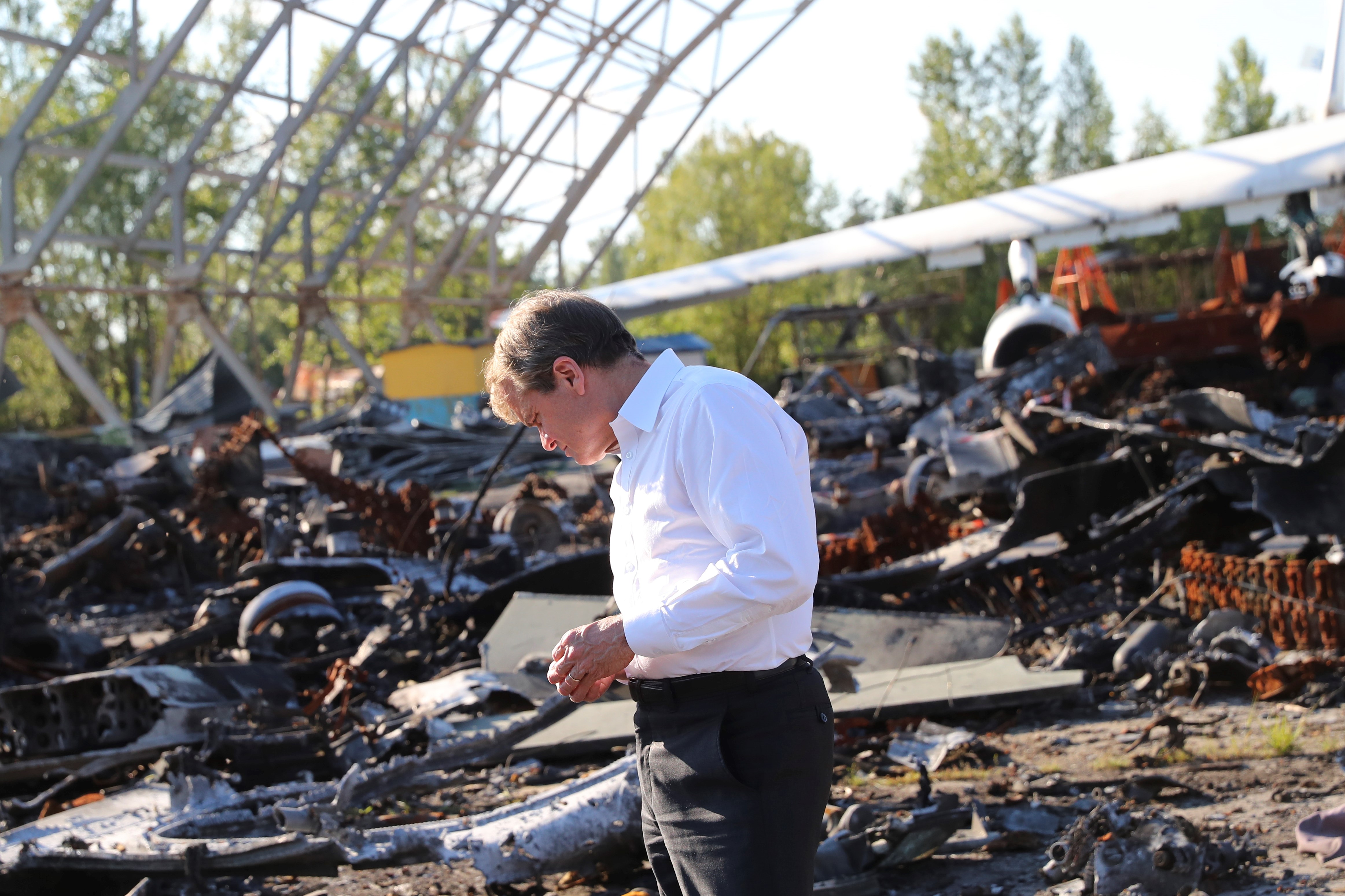 A man stands in black slacks and a white button down, looking somber, behind him are destroyed airplane parts and the remains of an airport hanger
