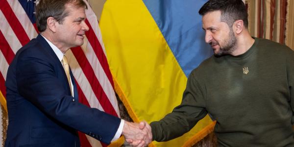 A man in a suit, Congressman Quigley, smiles while shaking hands with President Volodymyr Zelenskyy, who wears green military fatigues. They stand in front of US and Ukraine flags.