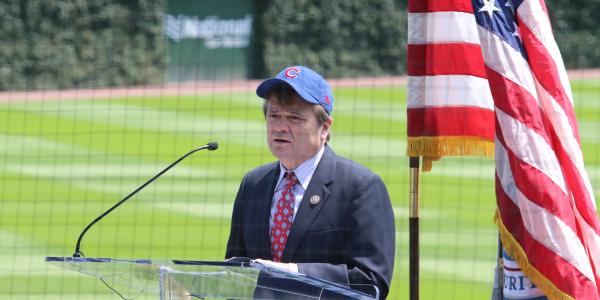 Quigley speaks from a podium with the American flag and green fields in the background. He wears a suit and blue Cubs baseball cap.