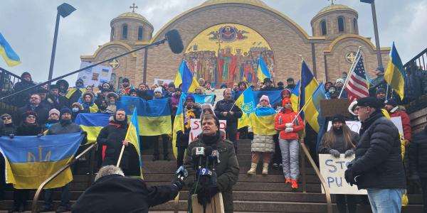 Mike Quigley speaks before microphones, behind him are rally  goers carrying Ukrainian flags