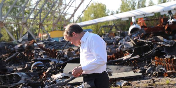 A man stands in black slacks and a white button down, looking somber, behind him are destroyed airplane parts and the remains of an airport hanger