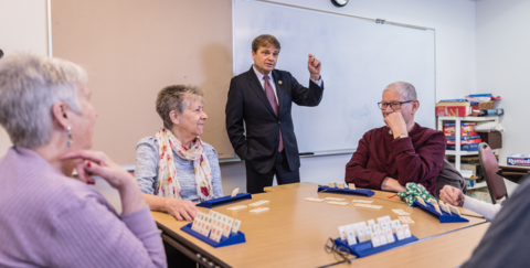 mike quigley stands in front of a table of people playing a card game. the players are all older.